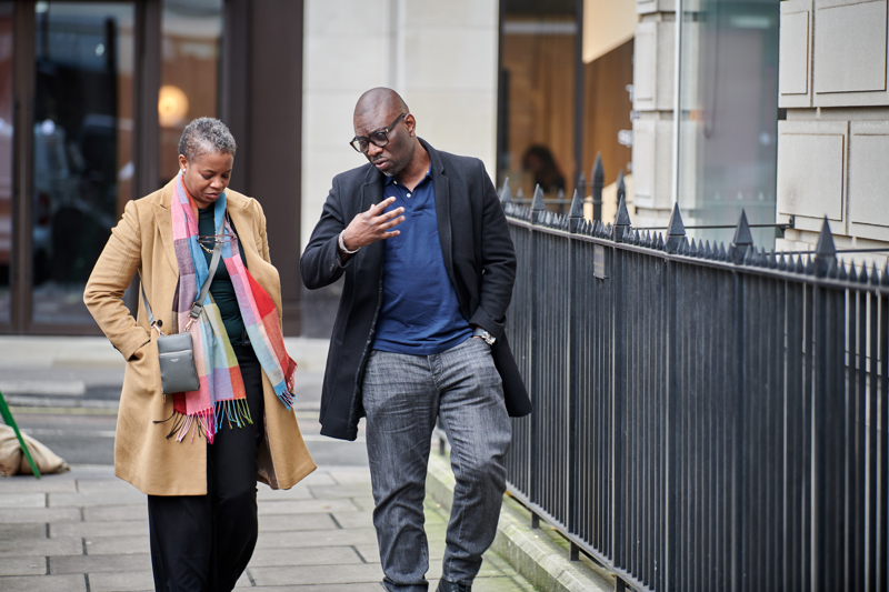 Black man in dark jeans with blue top and black coat and and woman in dark outfit with camel coat and multi-colour scarf walking through london street talking