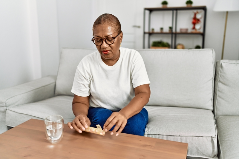 An older black female is taking a tablet out of a tablet organiser. She has a glass of water ready