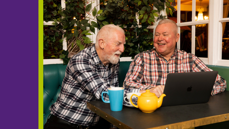 Two men, smiling, looking at an open laptop with a cup of tea.
